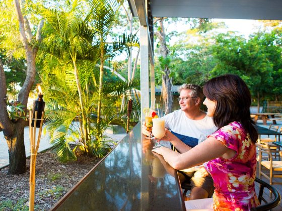 Two people sit at an outdoor bar counter surrounded by lush greenery, one holding a colourful drink with a straw and the other a light-coloured beverage, with palm trees, tiki torches, and warm sunlight creating a tropical atmosphere.