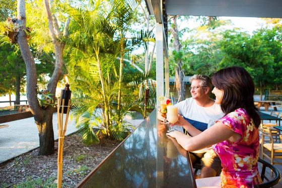 Two people sit at an outdoor bar counter surrounded by lush greenery, one holding a colourful drink with a straw and the other a light-coloured beverage, with palm trees, tiki torches, and warm sunlight creating a tropical atmosphere.