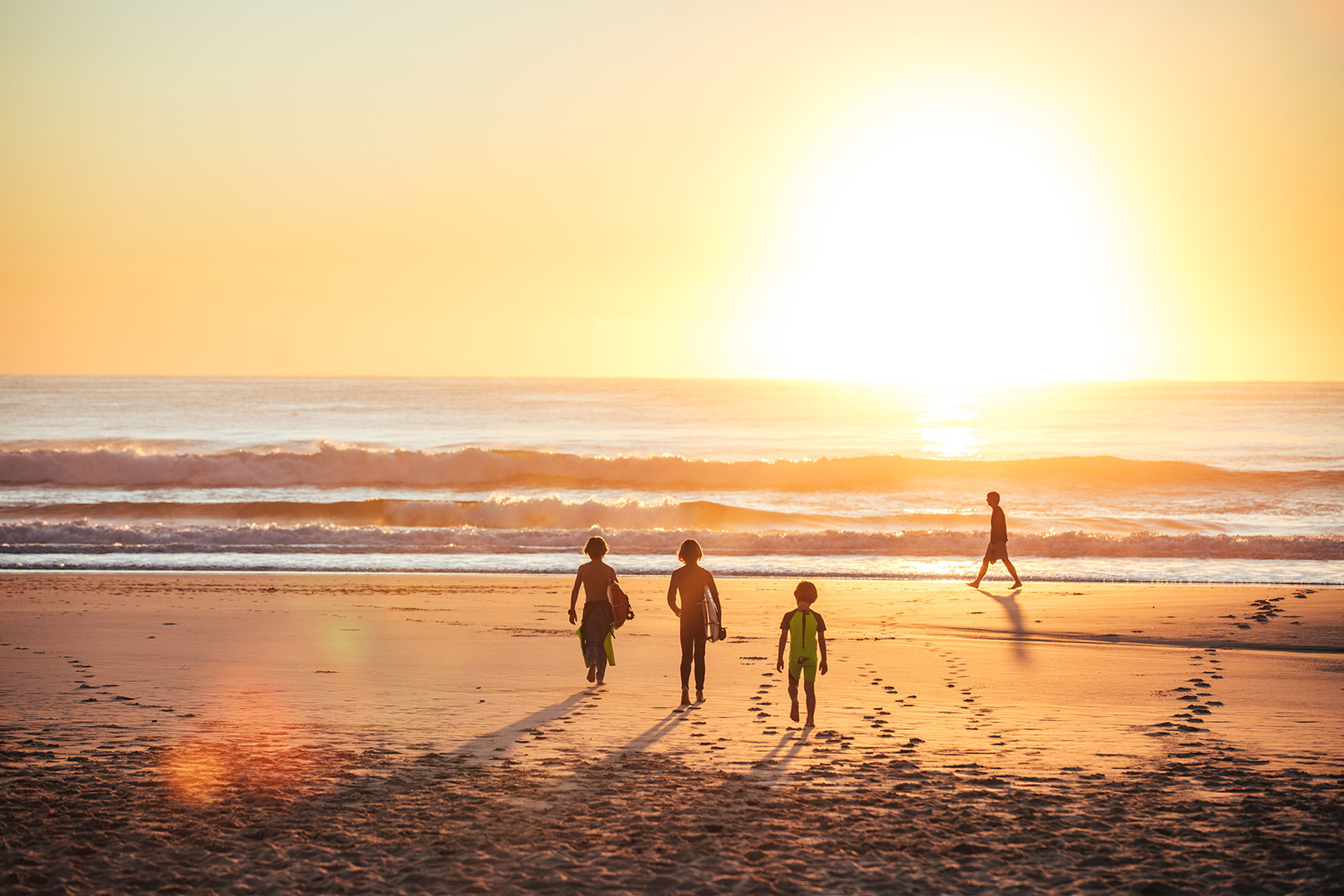 A group of young boys walking towards the beach with their surfboards. It's early morning and the sun is rising over the beach.