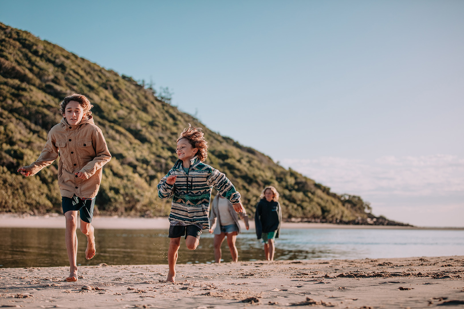 A young family running around on the sand at Tallebudgera Creek Beach. It is early morning and the family are wearing winter clothes.