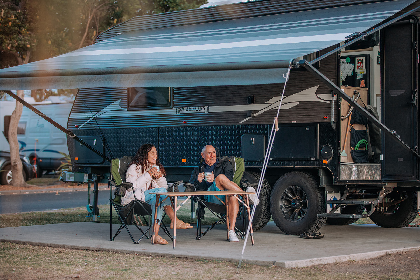 A middle aged couple sitting in camp chairs near the caravan. The are wearing winter jackets and sipping tea from mugs.