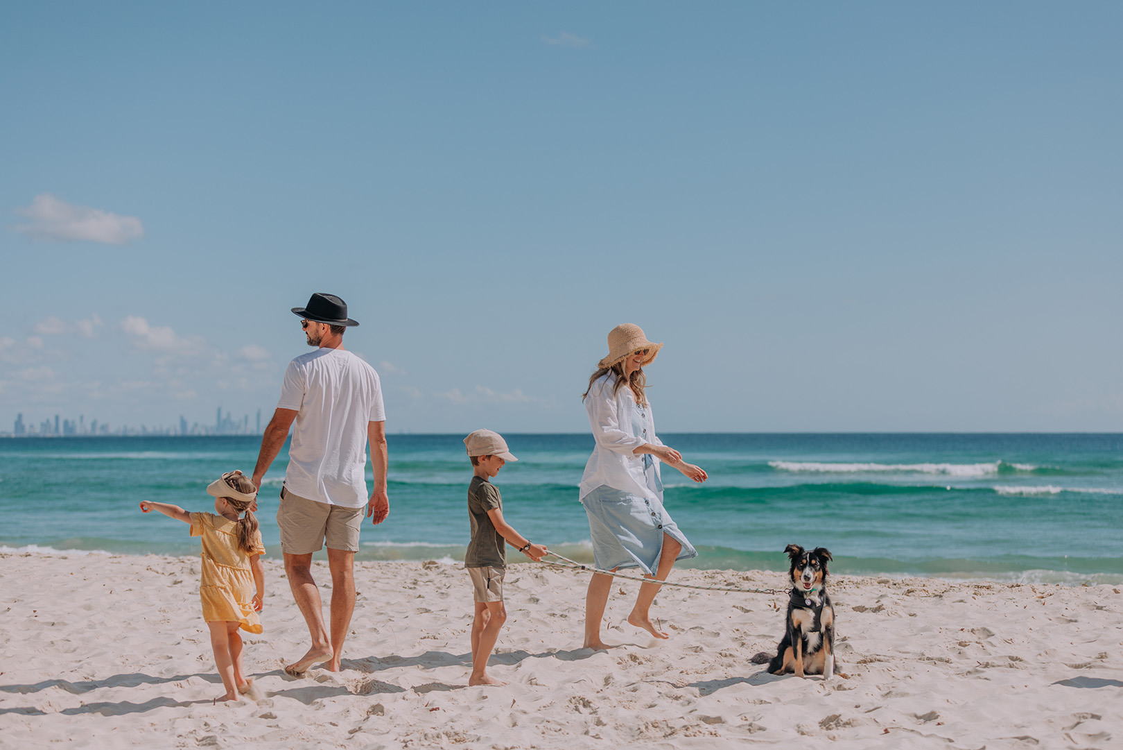 A family walking along the beach at Kirra with their dog. The sun is shining and the sky is blue.