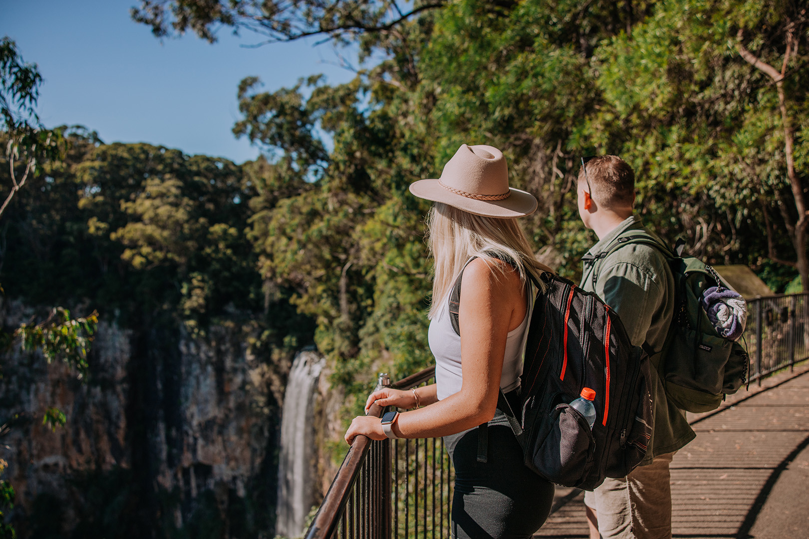 A couple standing at a lookout overlooking a large waterfall off a lush cliff. It's a clear sunny day.