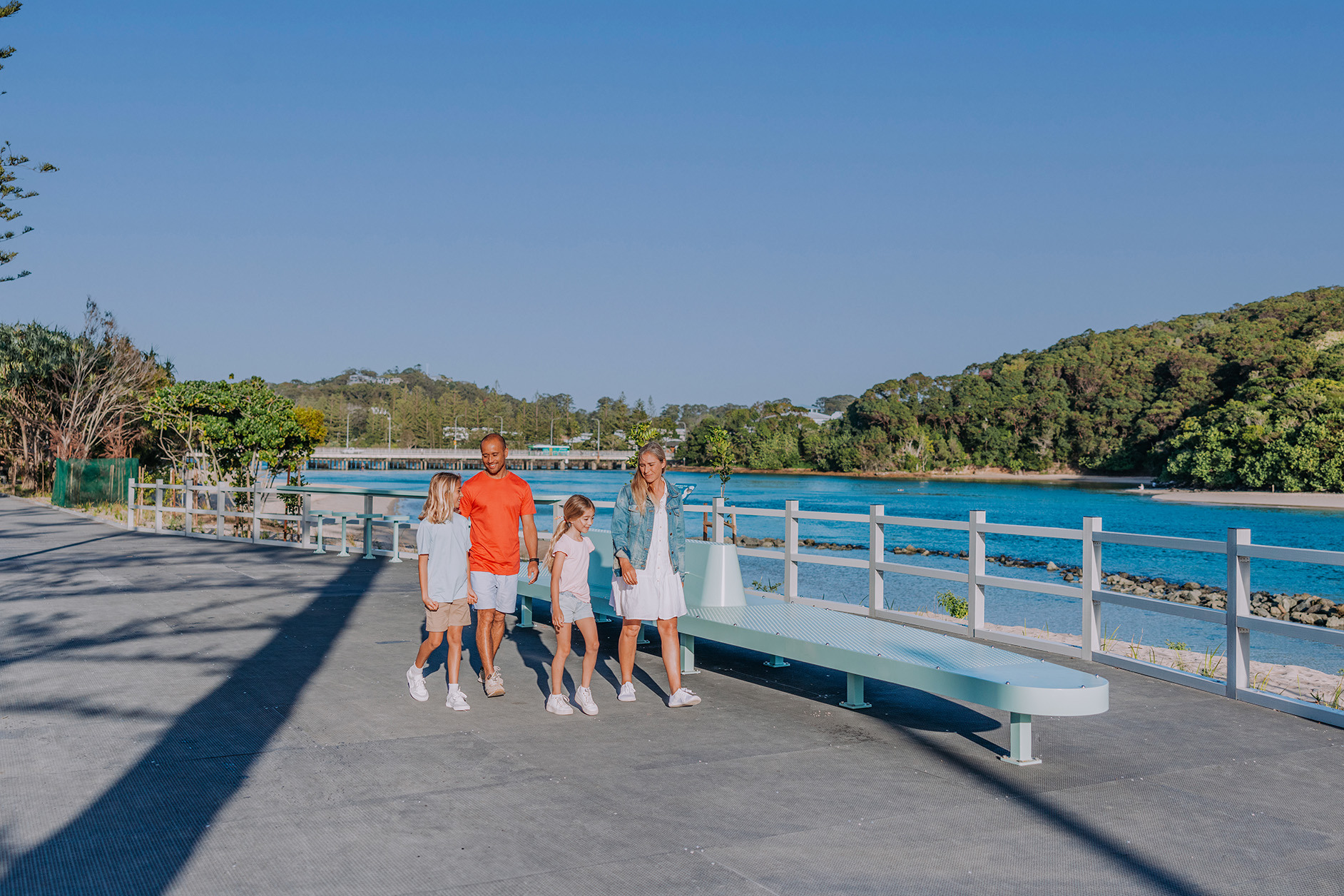 A family of four walk along a boardwalk at Tallebudgera Creek. The creek and the headland are in the distance.