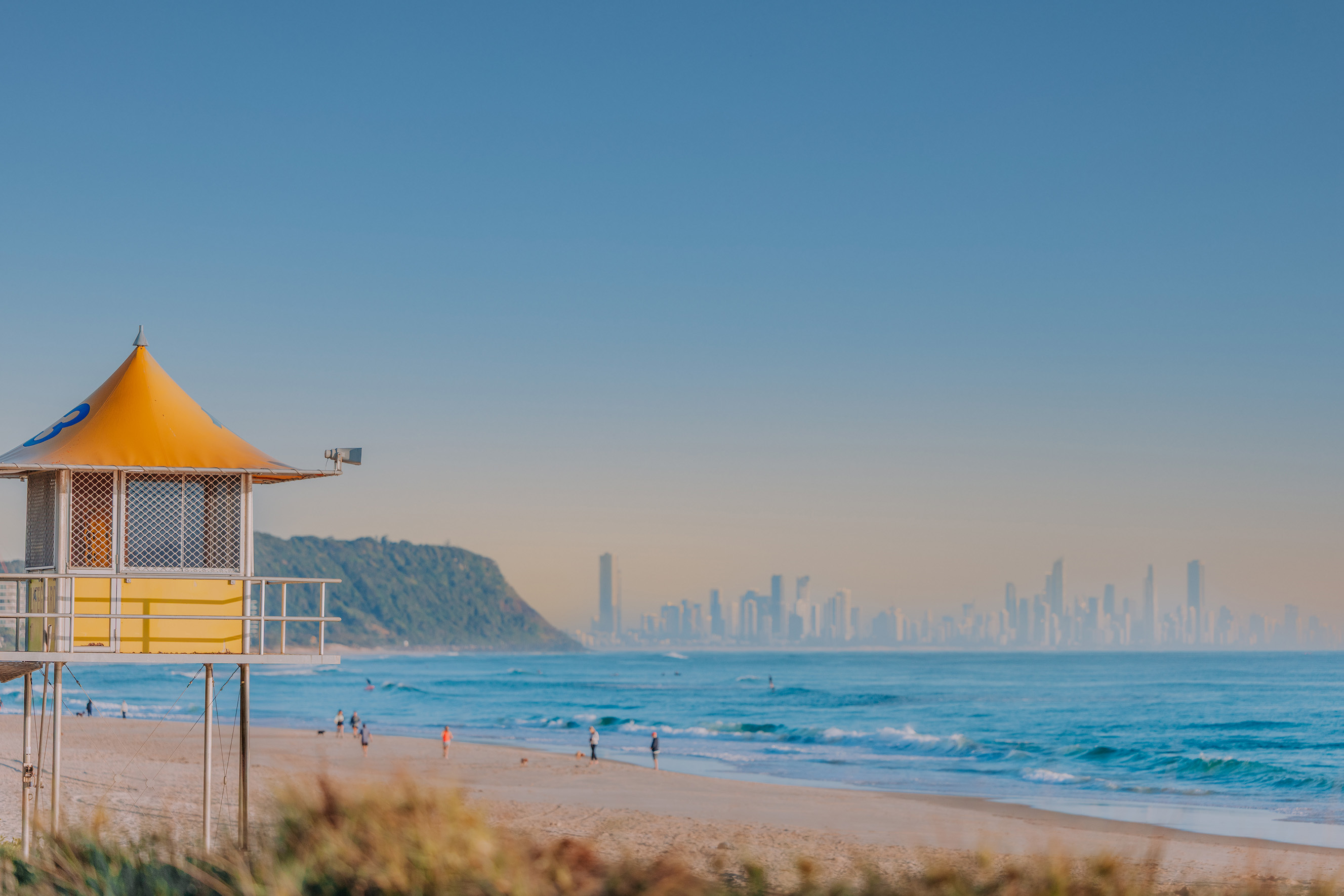 A lifeguard tower with a beautiful beach in the background at sunrise. People are walking along the beach and swimming.