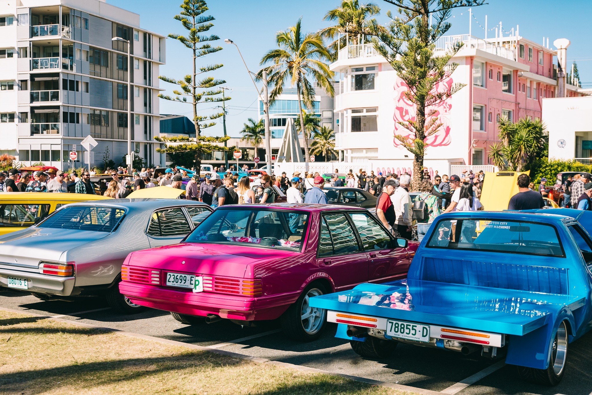 A row of colourful vintage cars at Cooly Rocks On festival in Coolangatta. The sun is shining and people are walking around looking at the cars.