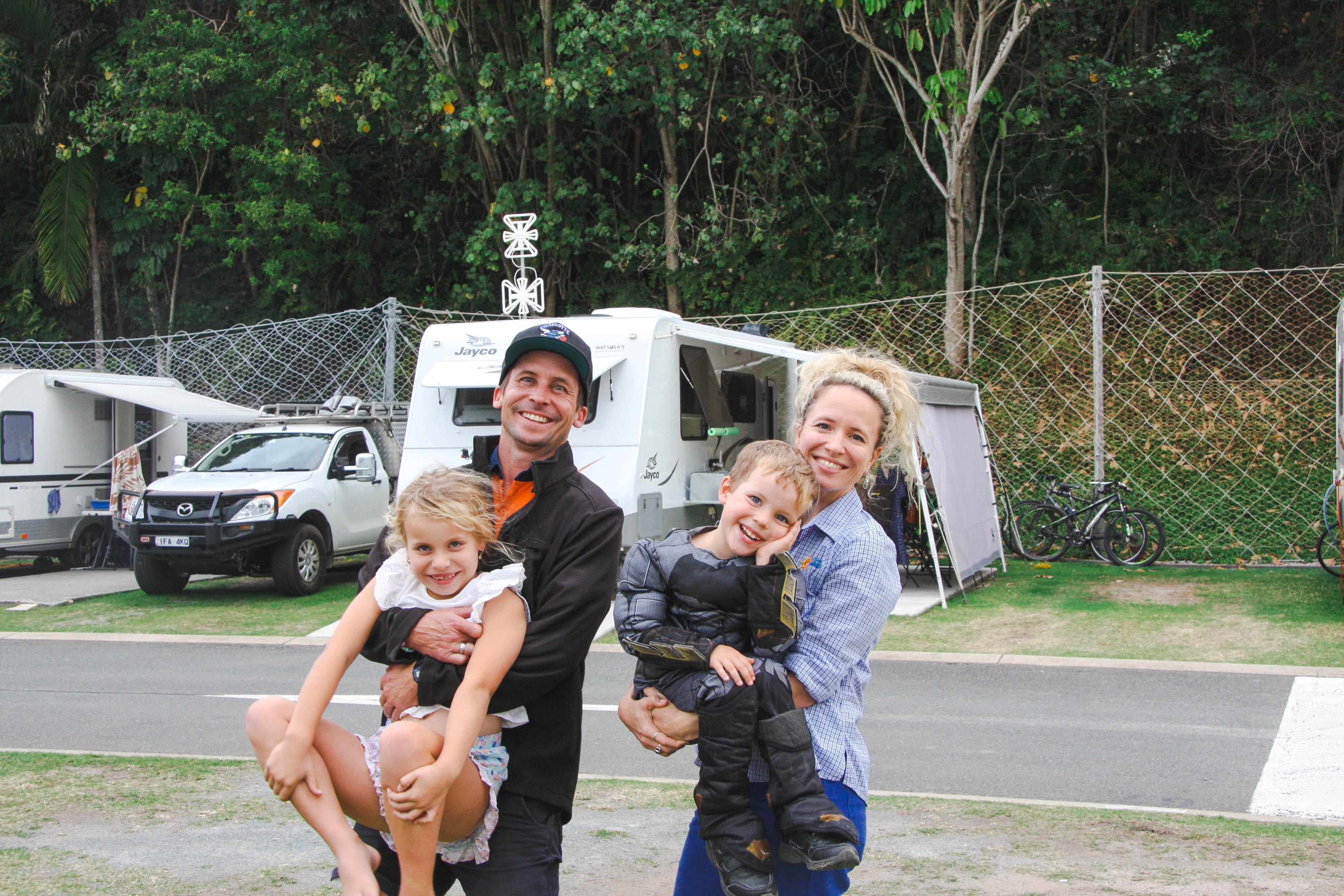 Park Manager of Ocean Beach Tourist Park, Becky and Pete, pose with their 2 young children in the park next to a caravan.