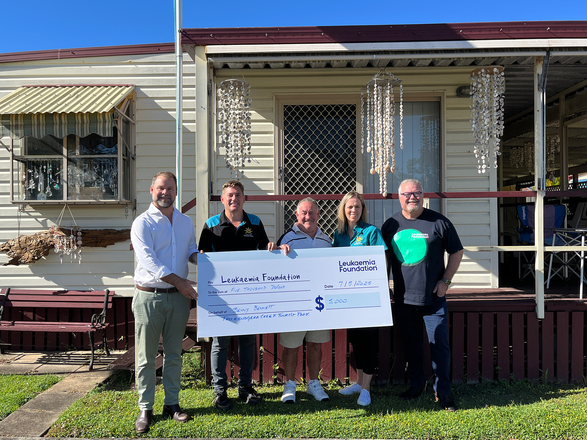 A group of five people standing in front of a house, holding a large ceremonial check from the Leukemia Foundation. The house has white siding with red trim and decorative hanging shells on the porch.