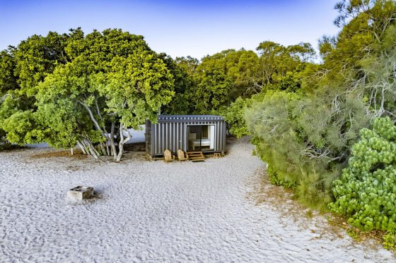 A small, modern cabin made from a shipping container sits on a sandy beach surrounded by lush green trees, featuring a wooden deck with two wicker chairs and a table, a nearby stone fire pit, and a clear blue sky overhead.