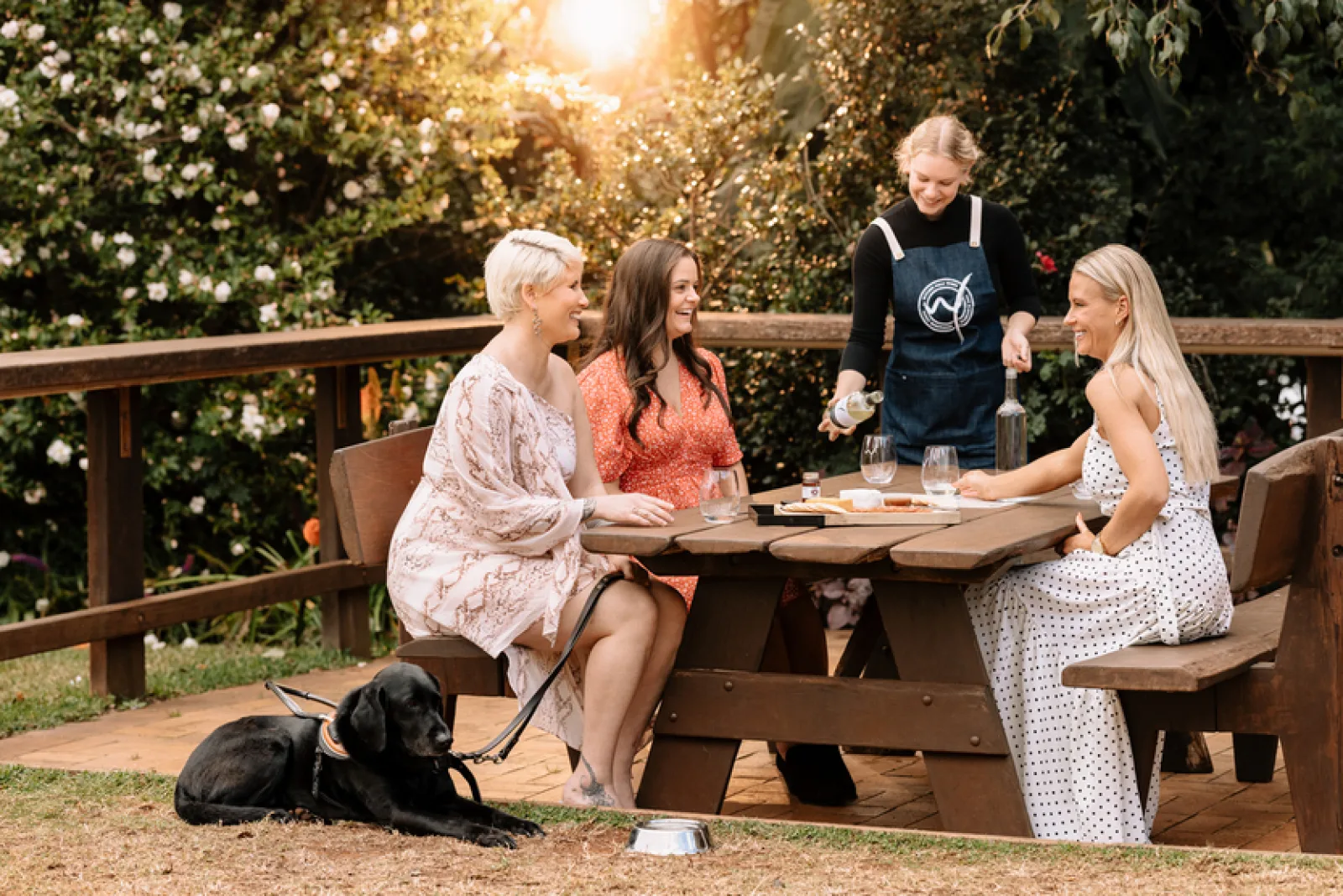 Three women sit at a picnic table where they are receiving a wine tasting.